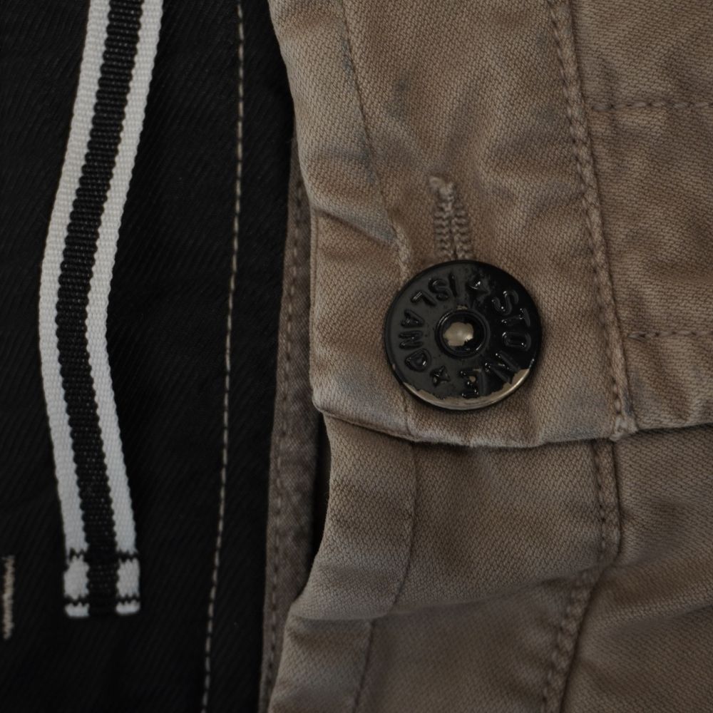 Primo piano del bottone nero in metallo logato Stone Island sulla chiusura dei pantaloni fango.