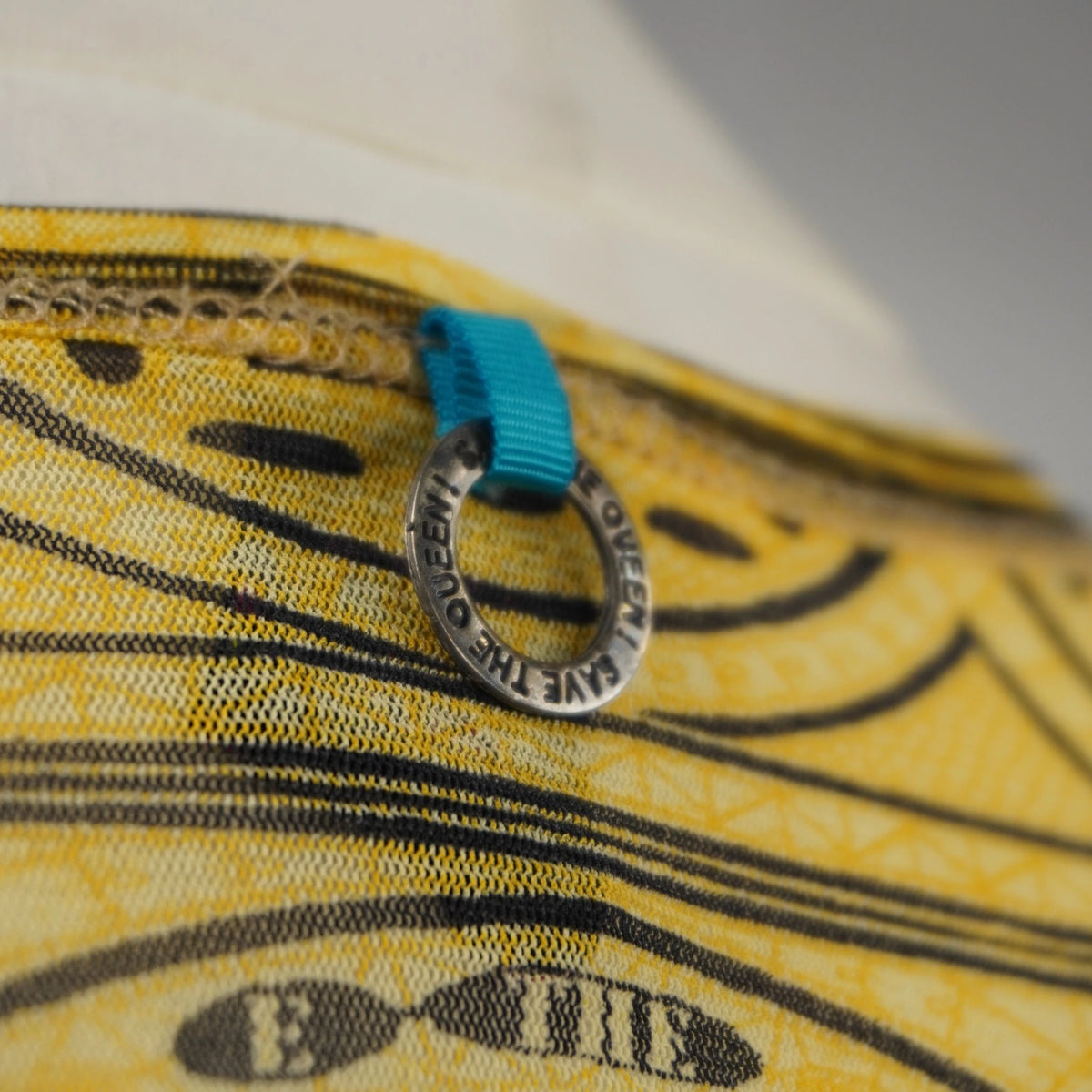 Macro del piccolo ciondolo circolare in metallo inciso con il brand Save The Queen! sul retro del collo.