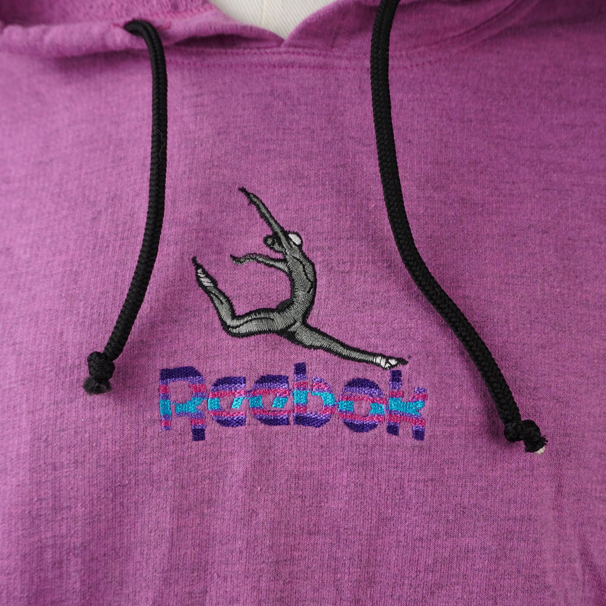 Primo piano del ricamo frontale multicolore Reebok con la figura di una ginnasta e cordini neri a contrasto.
