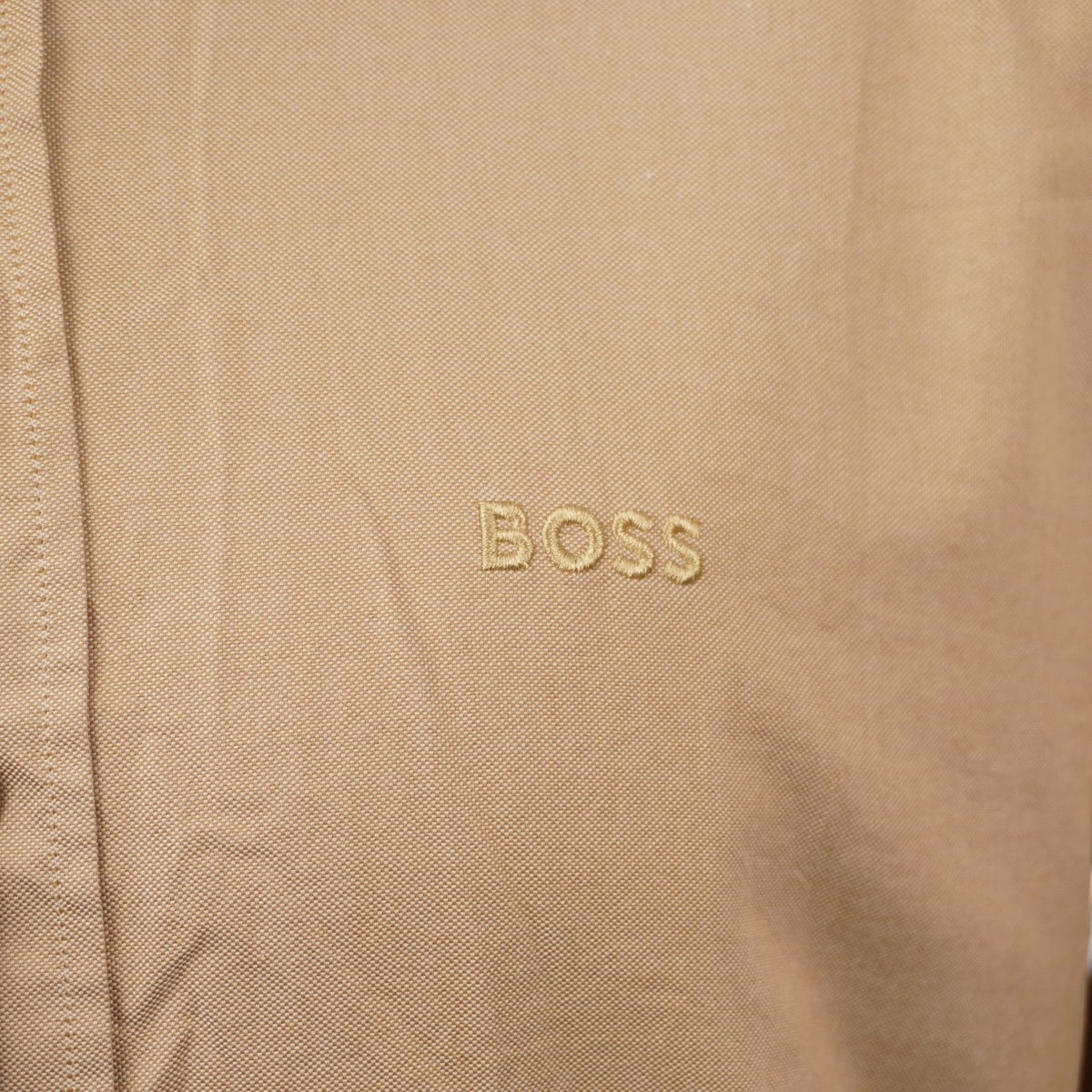 Inquadratura macro del tessuto in cotone organico beige che evidenzia il logo BOSS ricamato tono su tono sul petto.