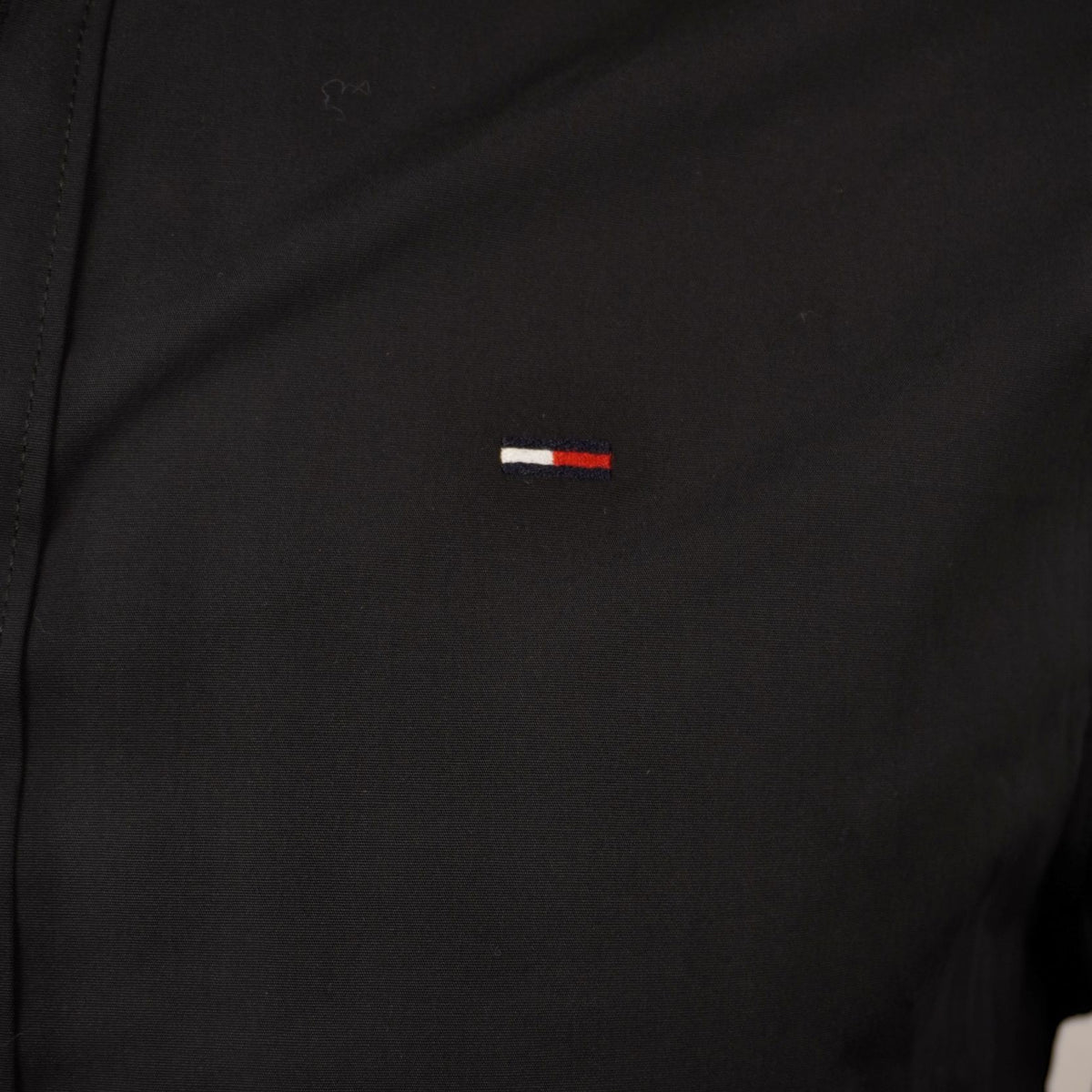 Inquadratura macro dell'iconico logo Flag di Tommy Jeans ricamato nei colori bianco, rosso e blu sul petto della camicia nera.